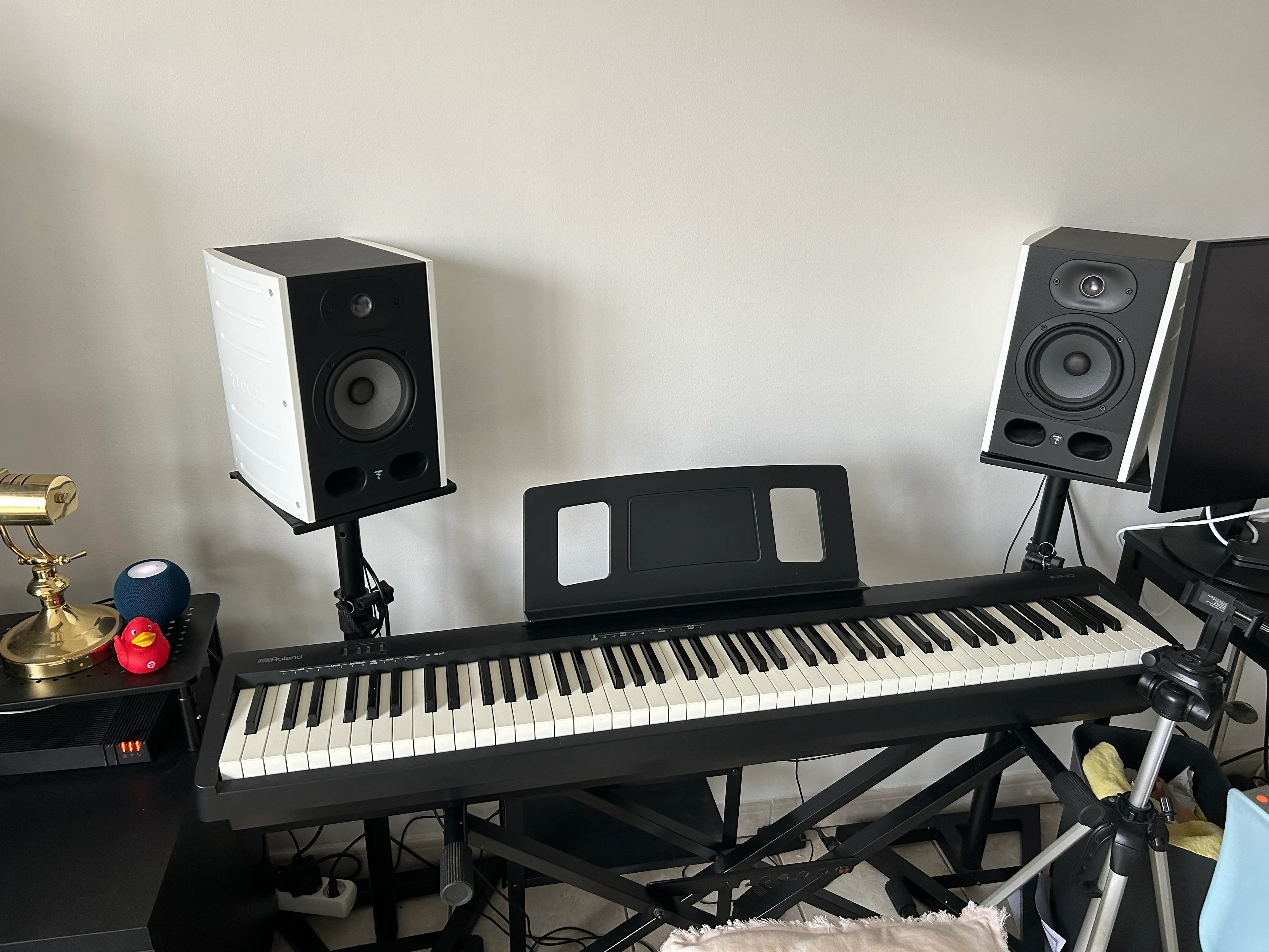 Un piano numérique et deux enceintes de la marque focale sur leurs pieds à 1m. Les enceintes sont de l'équipement de studio avec deux hauts parleurs chacun : un en métal et un en membrane. sur la gauche, un homepod et un canard en plastique rouge de Zenika, ainsi qu'une lampe de banquier couleur or.